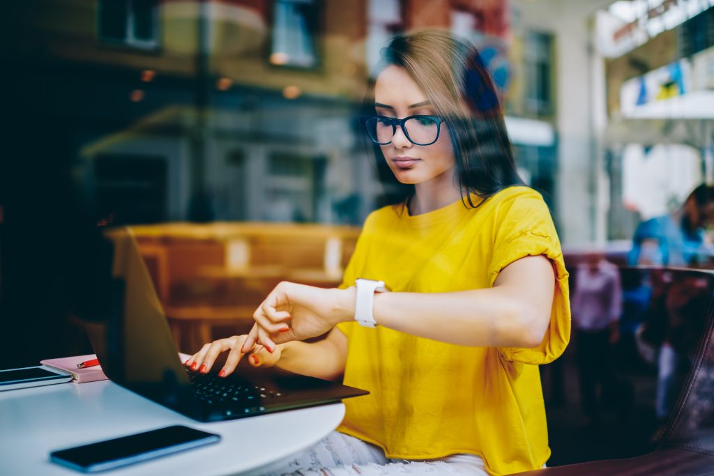 Weibliche Person mit braunen Haaren, Brille und gelben Shirt sitzt vor einem Laptop und schaut auf ihre Armbanduhr. Auf dem Tische vor ihr liegen zwei Telefone und ein Notizbuch.